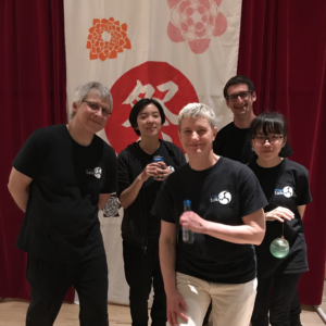 Five people in Pittsburgh Taiko t-shirts pose in front of the Matsuri banner. Some hold Ramune soda bottles or game prizes.